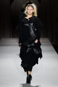 Model walking the runway in a black, layered outfit with leather panels and oversized coat, mid-stride on a white catwalk.