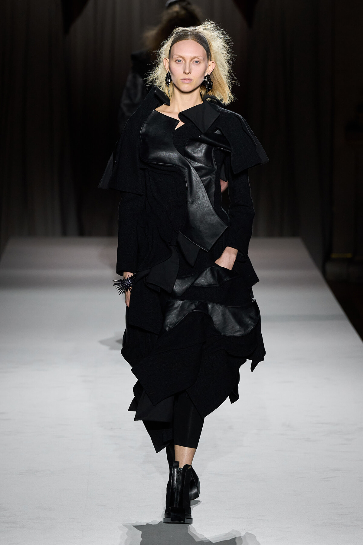 Model walking the runway in a black, layered outfit with leather panels and oversized coat, mid-stride on a white catwalk.