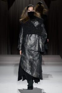 Model walking a fashion runway in a metallic gray coat layered over a black turtleneck and speckled cropped vest, face masked for the look.