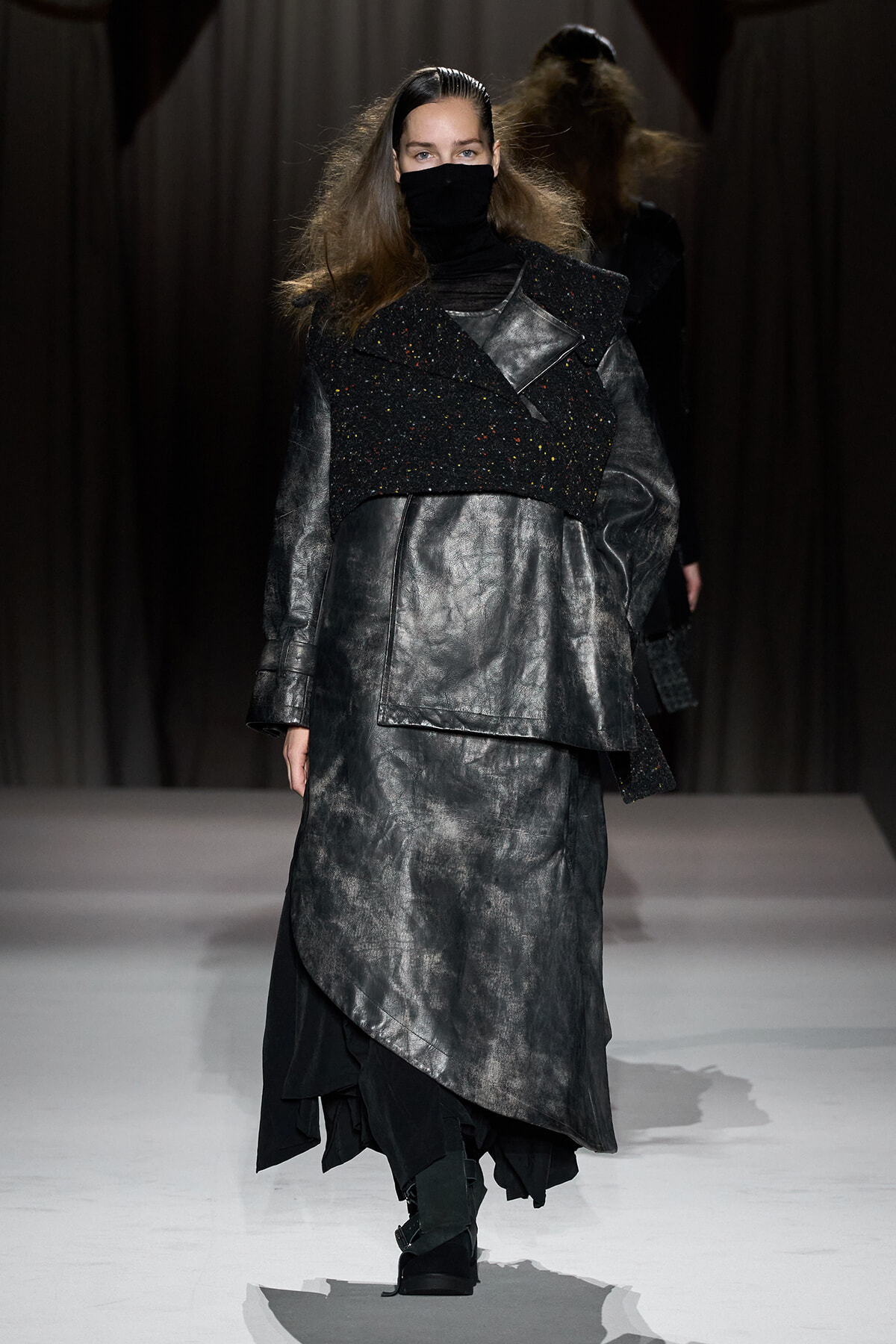 Model walking a fashion runway in a metallic gray coat layered over a black turtleneck and speckled cropped vest, face masked for the look.