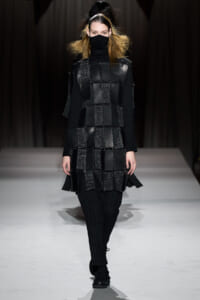 Model on a runway wearing a black turtleneck/ski-mask, a patchwork square-panel tunic made of glossy black fabrics, and black pinstripe pants with chunky shoes.
