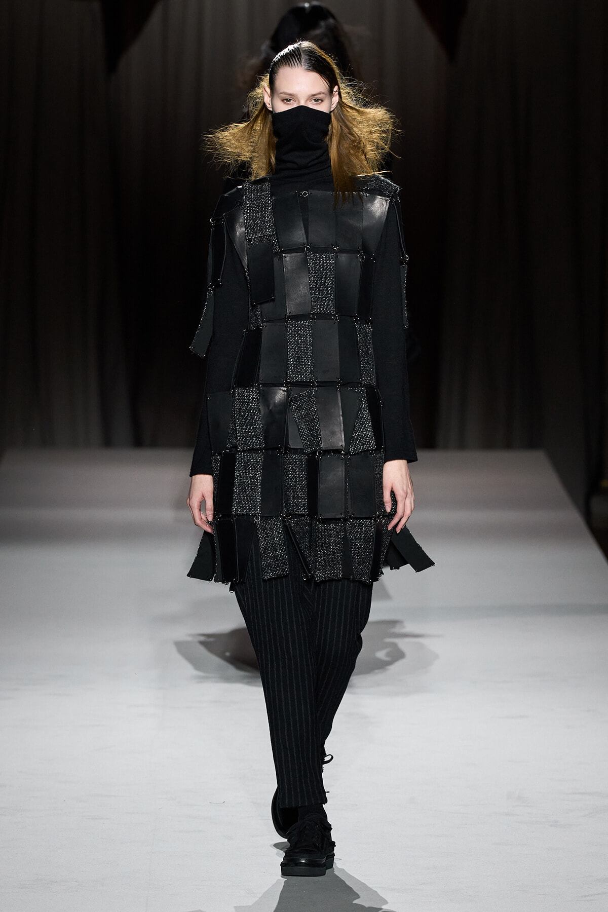 Model on a runway wearing a black turtleneck/ski-mask, a patchwork square-panel tunic made of glossy black fabrics, and black pinstripe pants with chunky shoes.