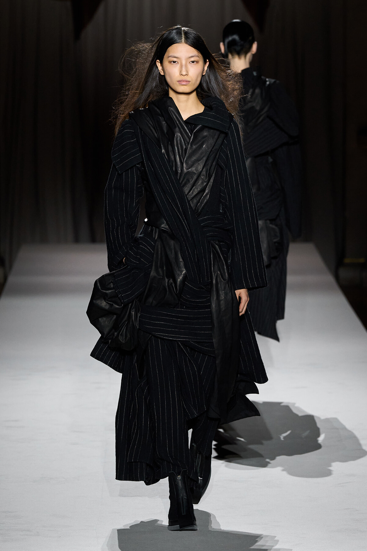 Model walking the runway in a black pinstripe outfit with layered, draped fabric and ankle boots.
