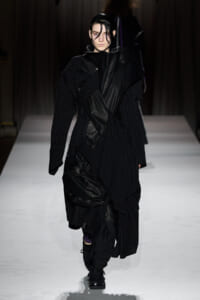 Model walking a runway in a dramatic black layered coat with draped fabrics and pinstripe suit elements, dark wet hair, serious expression.