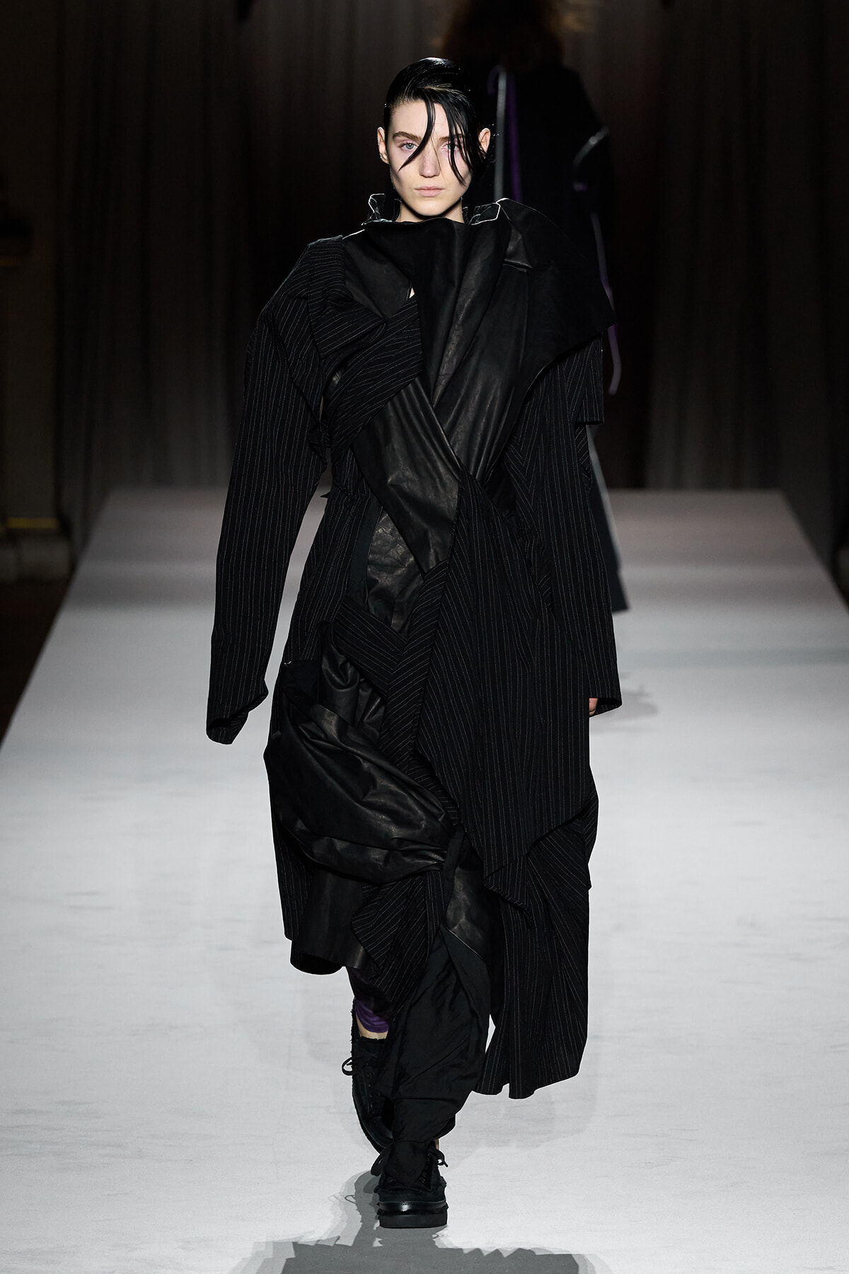 Model walking a runway in a dramatic black layered coat with draped fabrics and pinstripe suit elements, dark wet hair, serious expression.