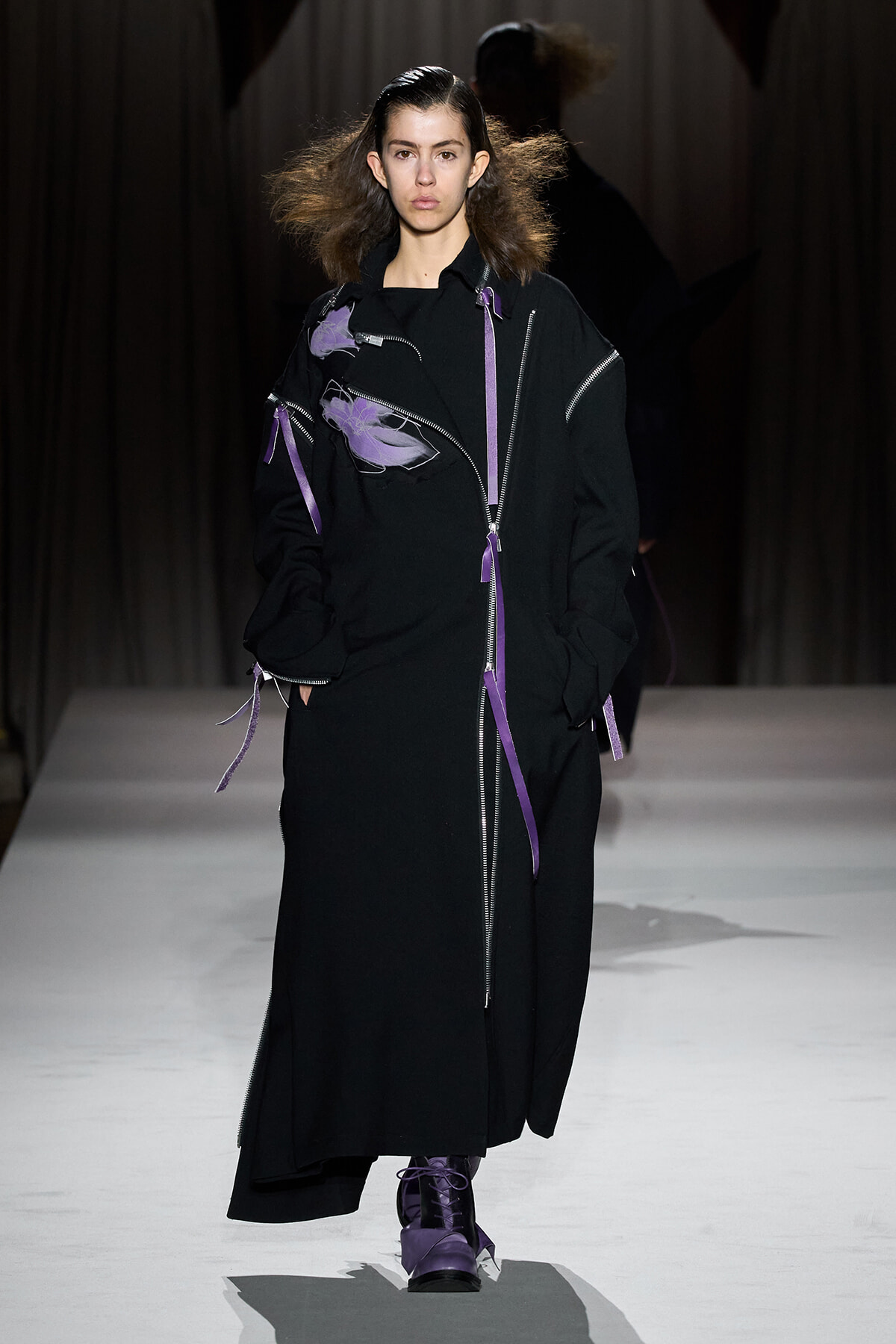 Model walking a fashion runway in a long black coat with purple zippers and ribbons, paired with purple boots.",