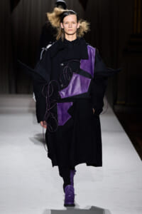 Model walks the runway in a long black coat with purple leather patches and purple cords draped across the front, paired with purple boots.