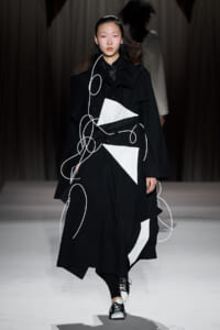 Model walks the runway in a black trench with white geometric panels and looping white cords.