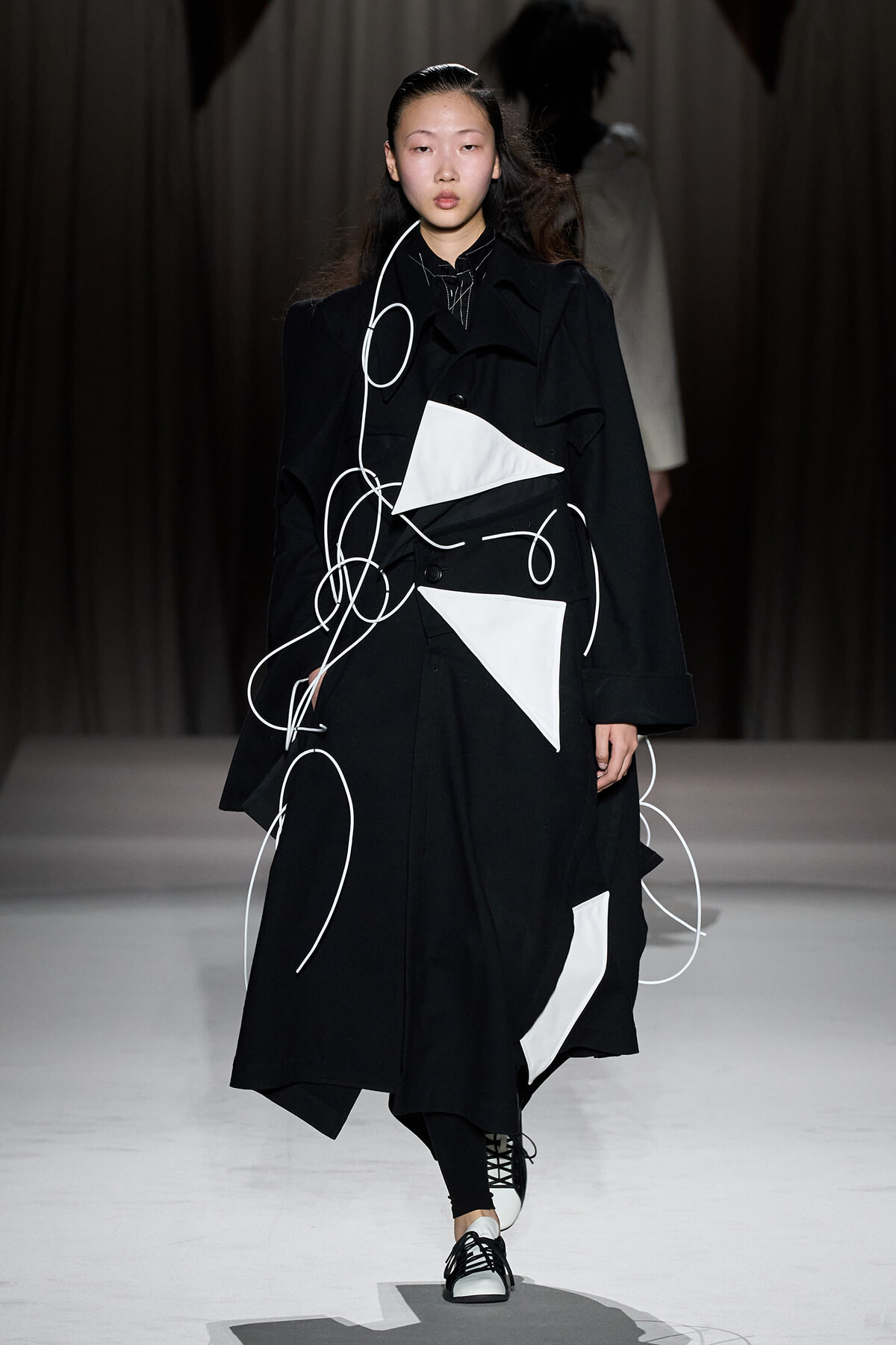 Model walks the runway in a black trench with white geometric panels and looping white cords.