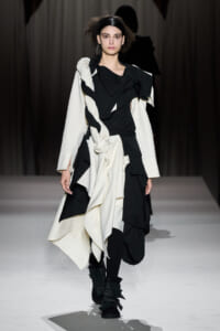 Model walking the runway in a black and white layered dress with oversized white coat and wrapped fabric details.