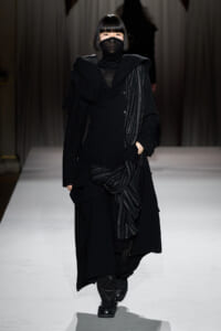 Model walks down a runway in an all-black, layered outfit with a knotted scarf and face mask.