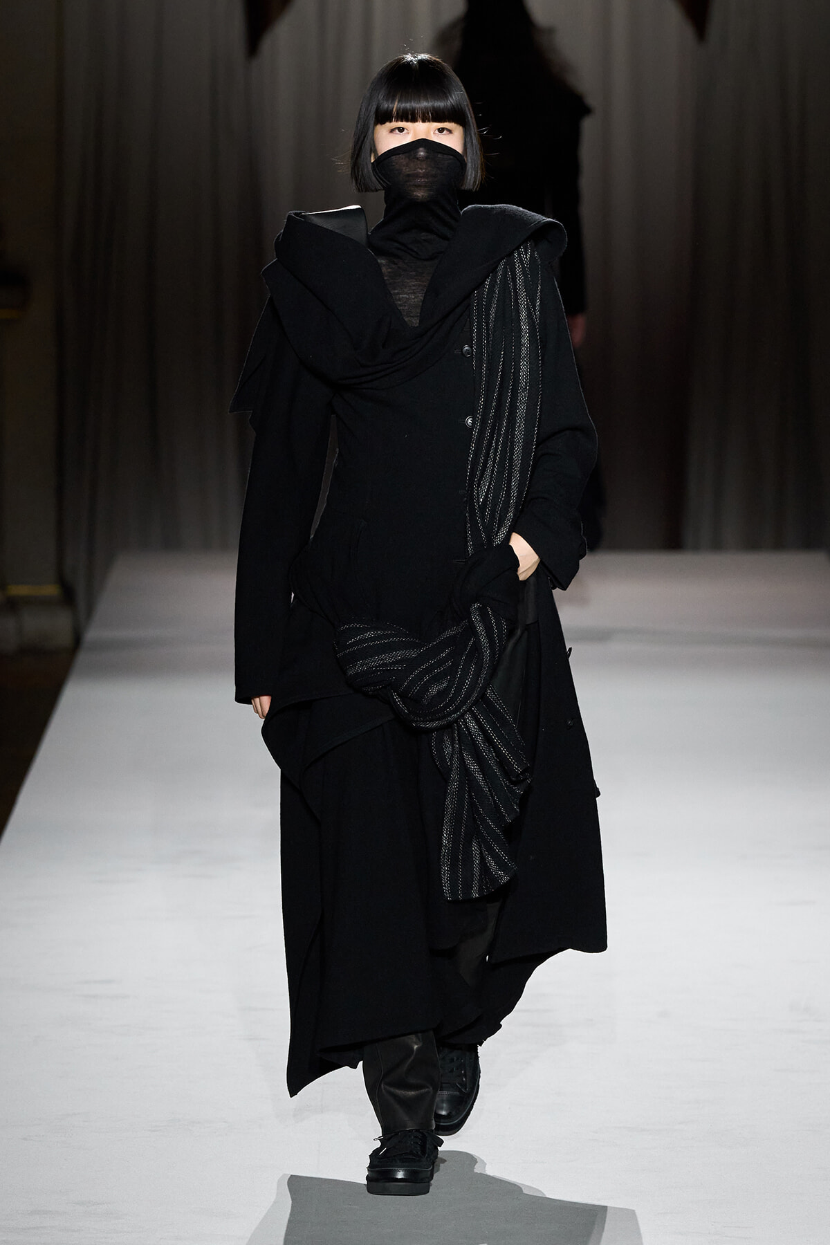 Model walks down a runway in an all-black, layered outfit with a knotted scarf and face mask.
