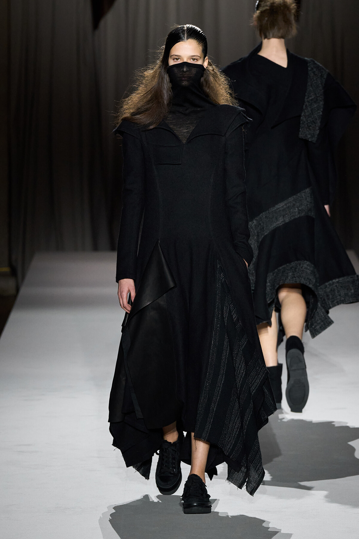 Model walks the runway in an all-black, layered outfit with a high neck and face covering, on a white catwalk.
