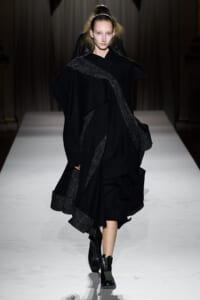 Model walking the runway in an all-black, oversized cloak with silver metallic trim on a white catwalk. Boots complete the look.