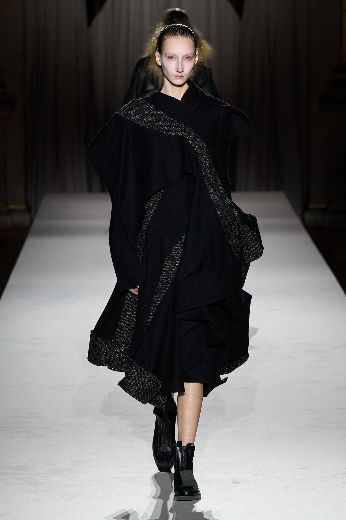 Model walking the runway in an all-black, oversized cloak with silver metallic trim on a white catwalk. Boots complete the look.