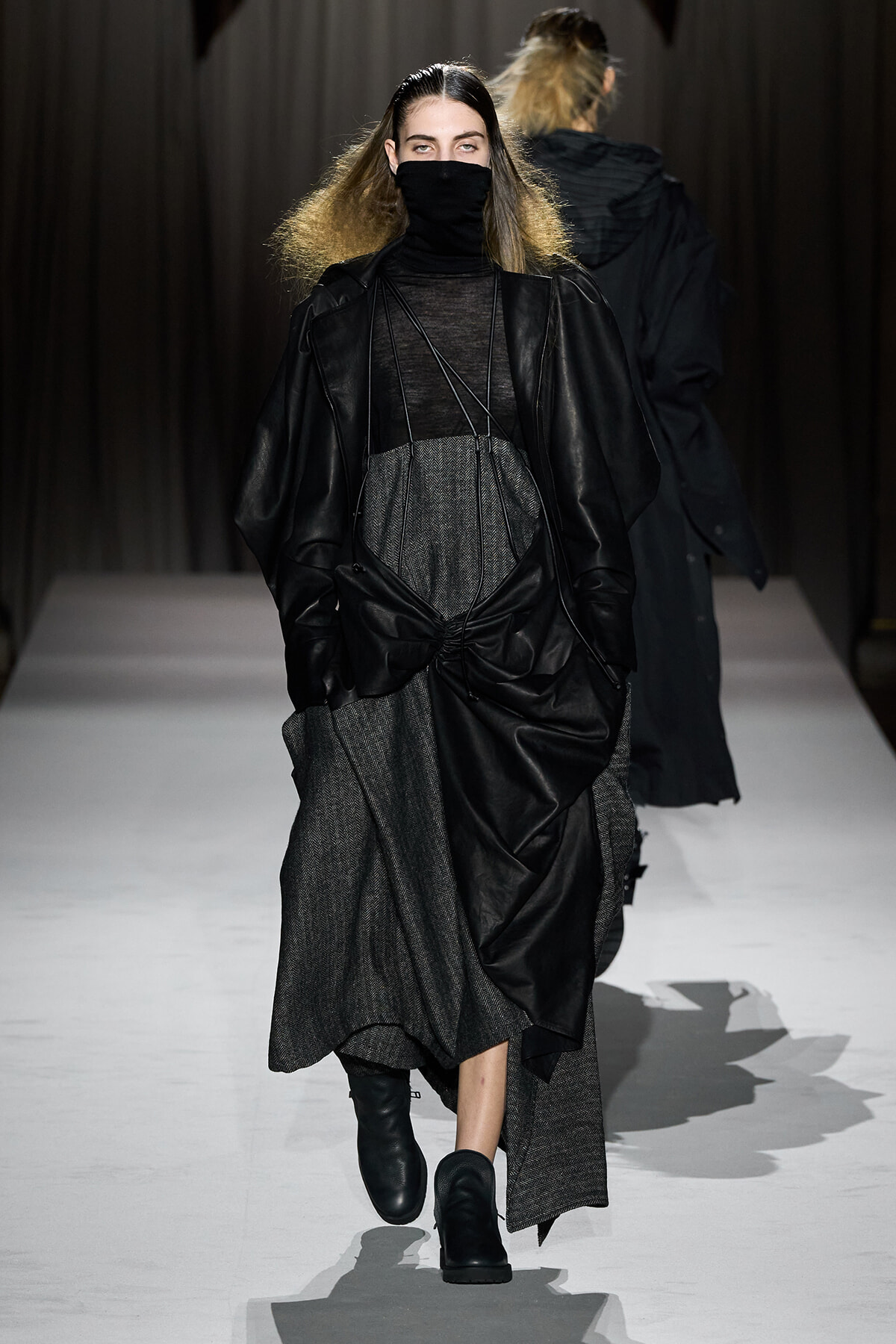 Model walking a runway in a black leather coat over a sheer top and gray textured dress, with a black face mask.