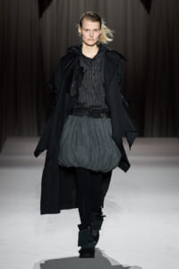 Model walking a fashion runway in a black trench, striped hoodie, and oversized gray bubble skirt with black tights and chunky boots