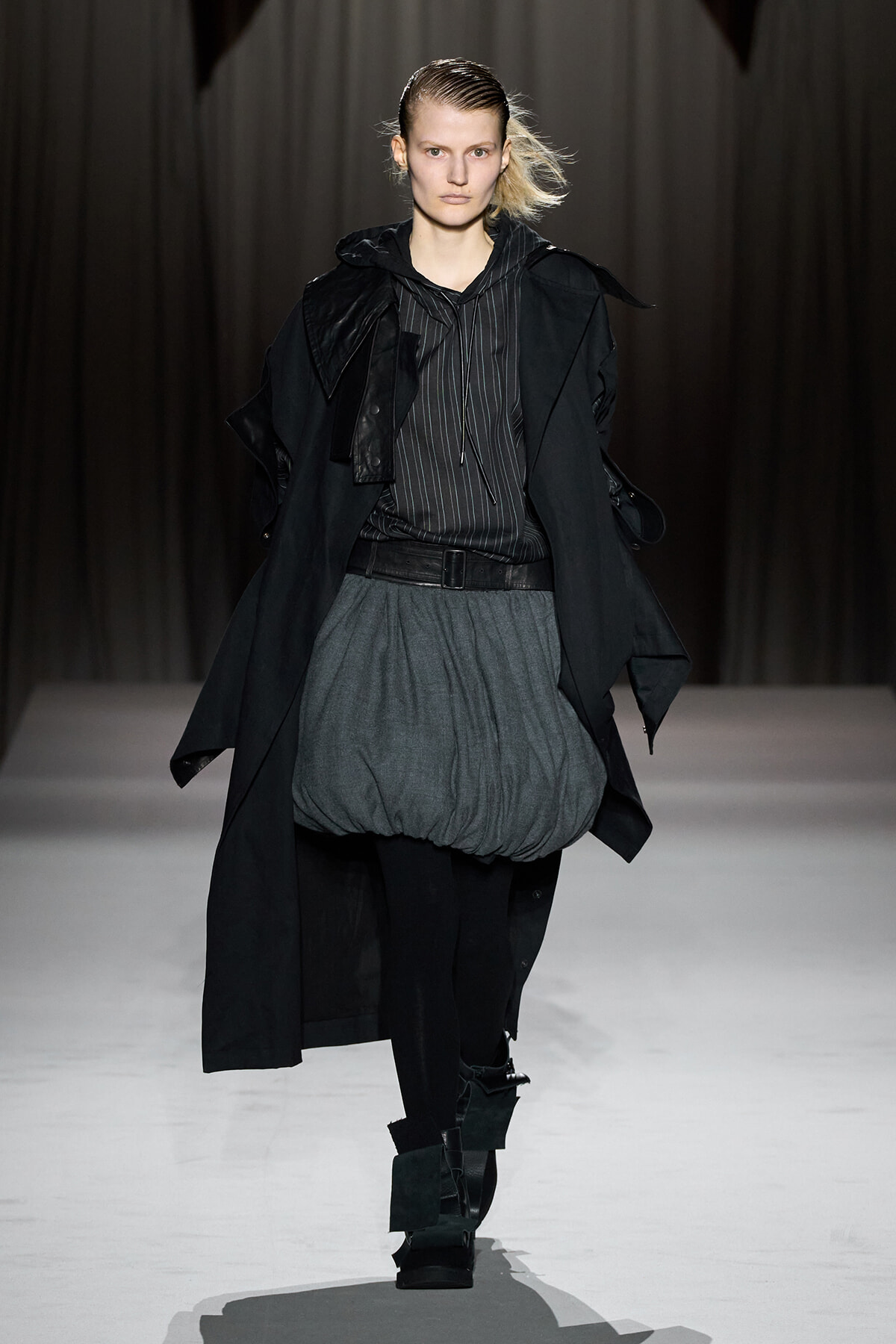 Model walking a fashion runway in a black trench, striped hoodie, and oversized gray bubble skirt with black tights and chunky boots