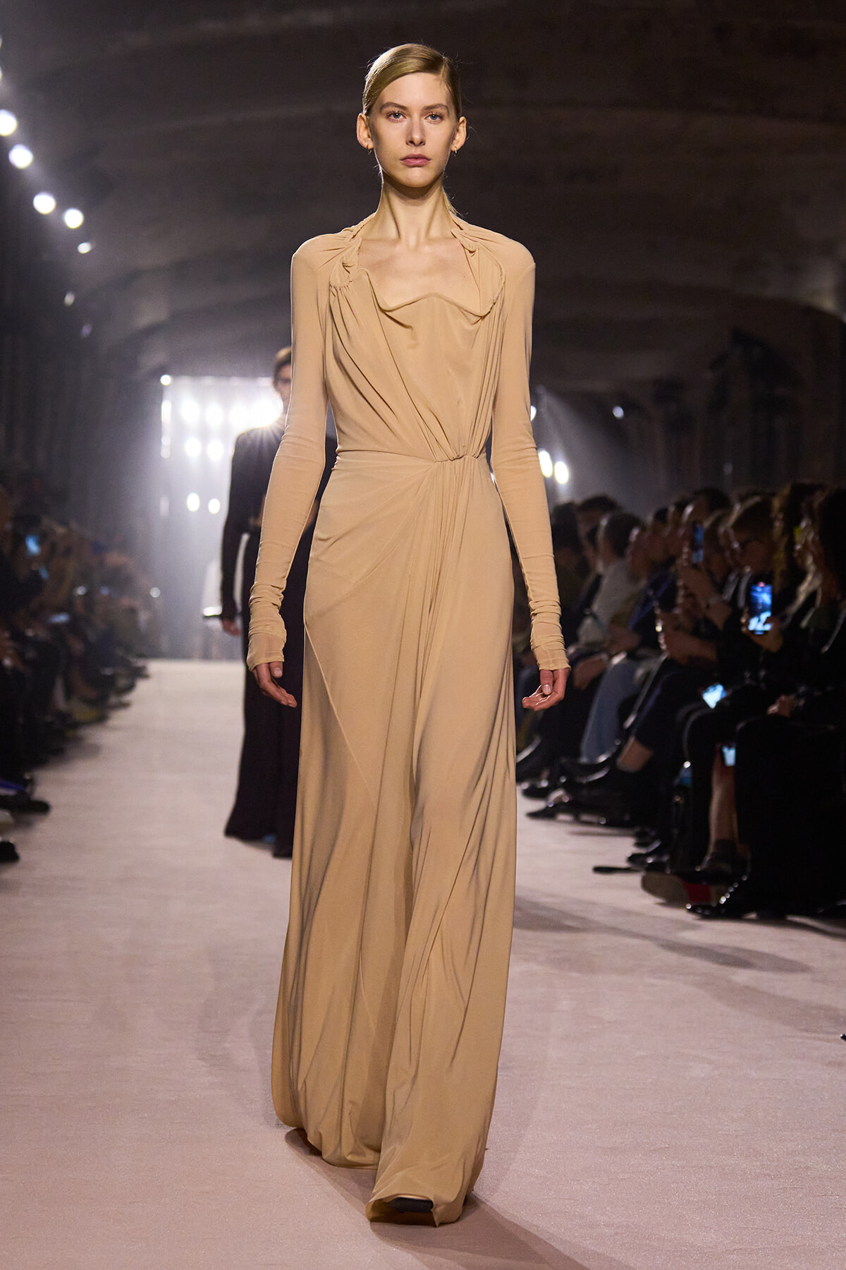 Model in a floor-length beige draped gown walks the runway during a fashion show, with seated audience on both sides illuminated by spotlights.