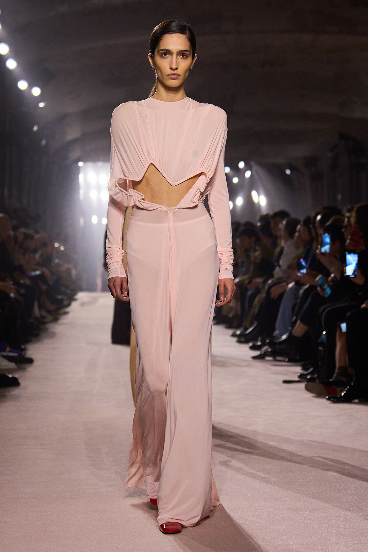 Model walks the runway in a pale pink, draped two-piece top with a midriff cutout and matching floor-length skirt, pink footwear visible, backstage lights in background.