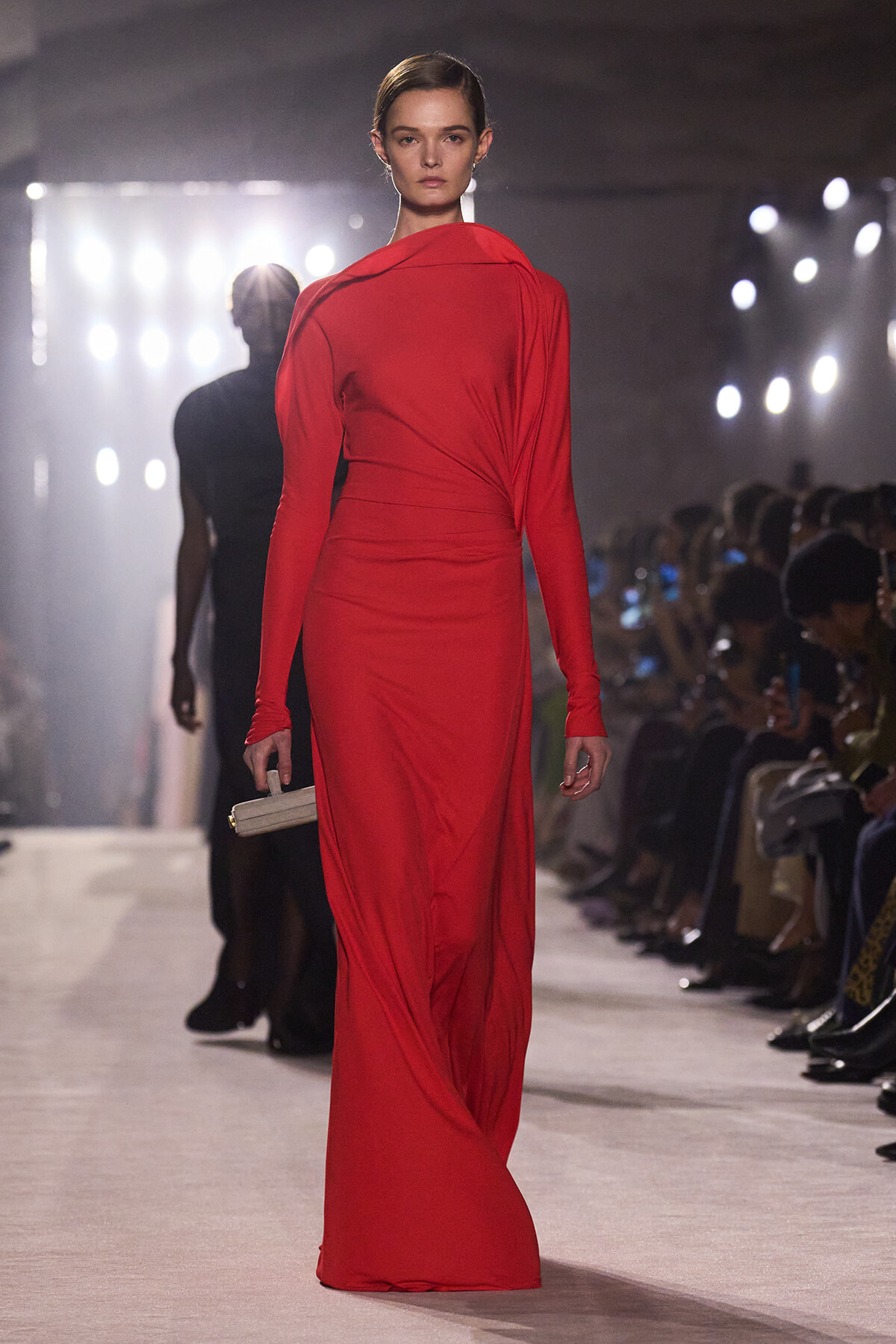 Model walks the runway in a red, floor-length, long-sleeve gown with a wrapped waist, holding a small clutch.