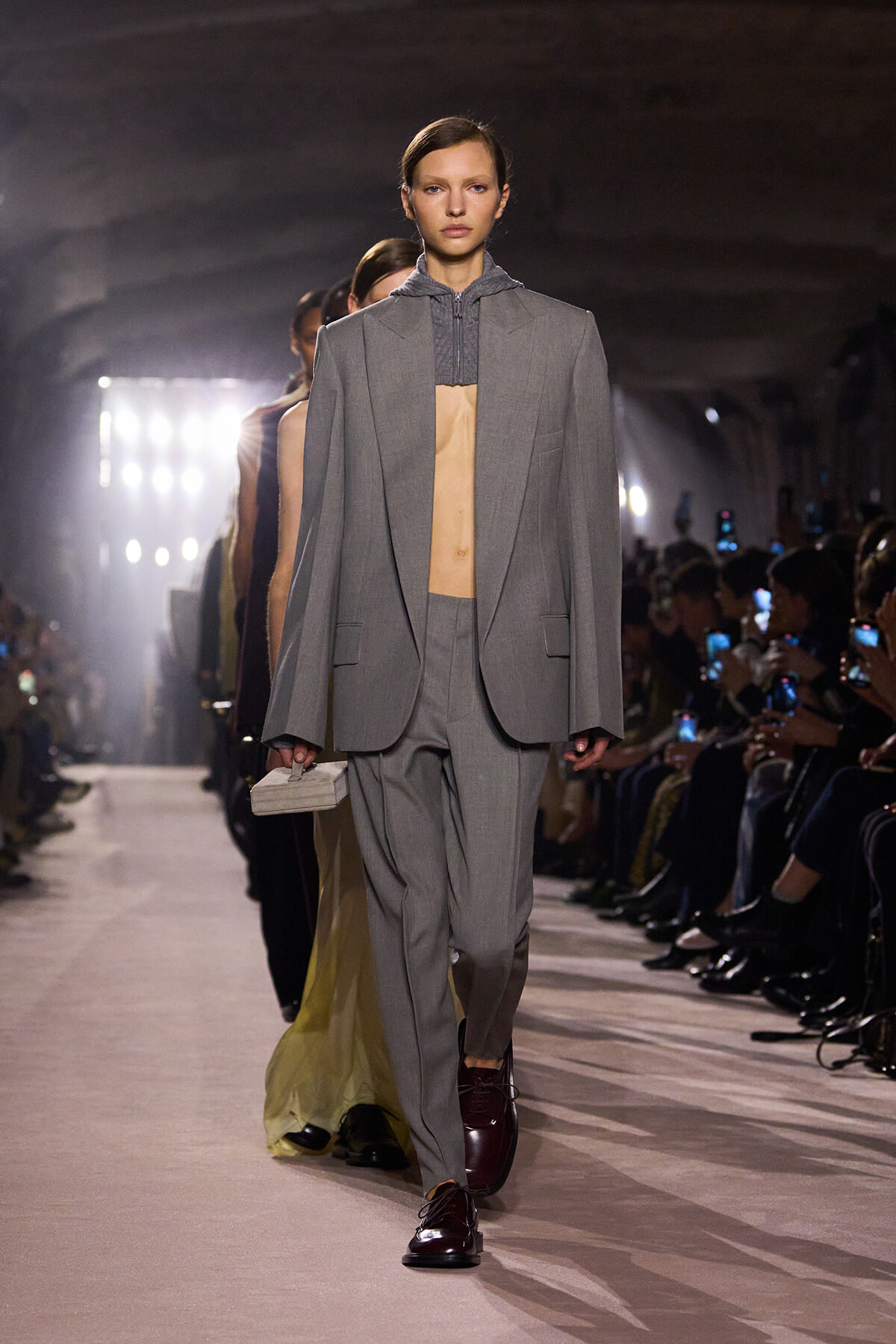 Runway model walking in a light gray oversized suit with matching blazer and trousers, shown with burgundy shoes and a small rectangular clutch in hand.