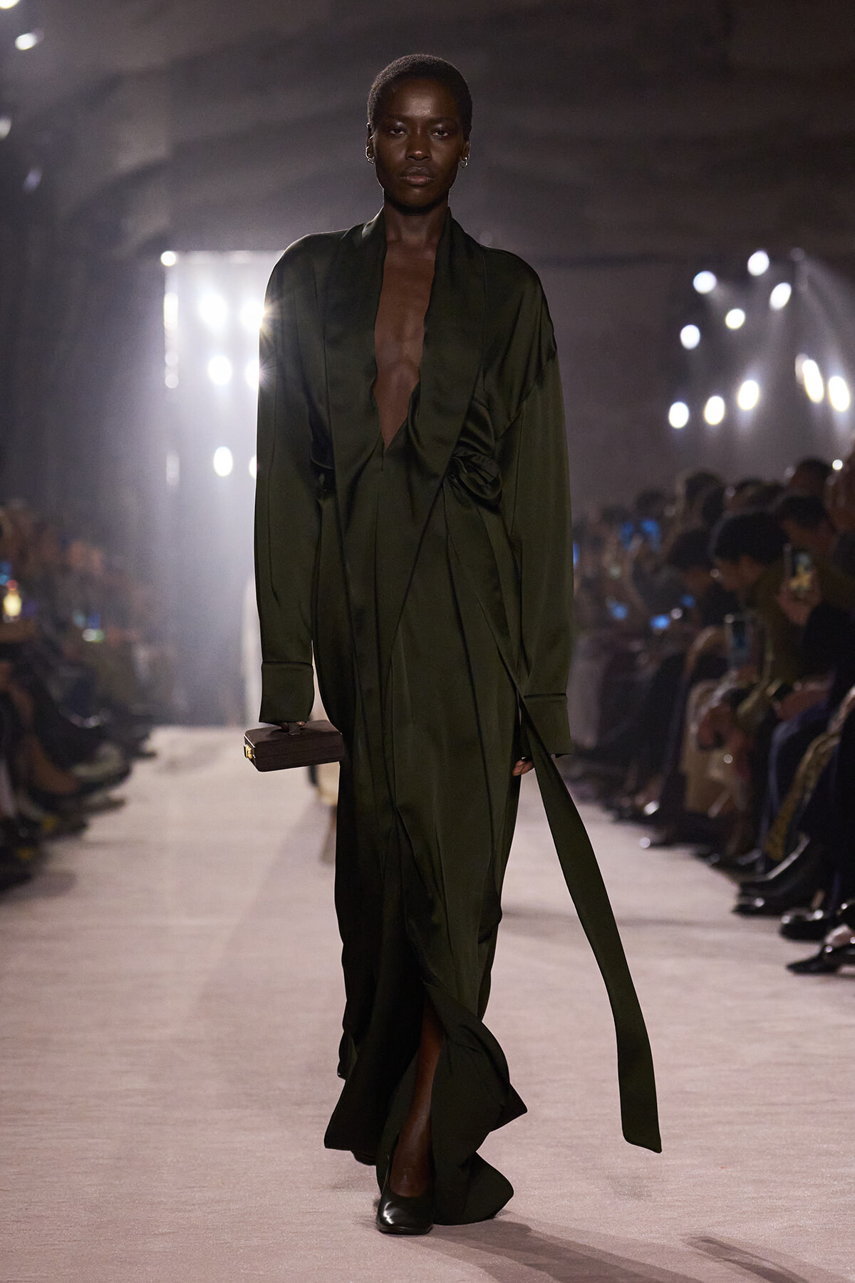 Model walks the runway in a deep olive-green draped wrap dress with a plunging neckline, holding a small rectangular clutch; dim backstage lighting and an audience in the background.