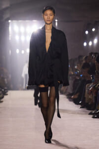 Model walking a fashion runway in a loose black wrap blouse, black shorts, sheer tights, and black heels, carrying a small rectangular clutch.