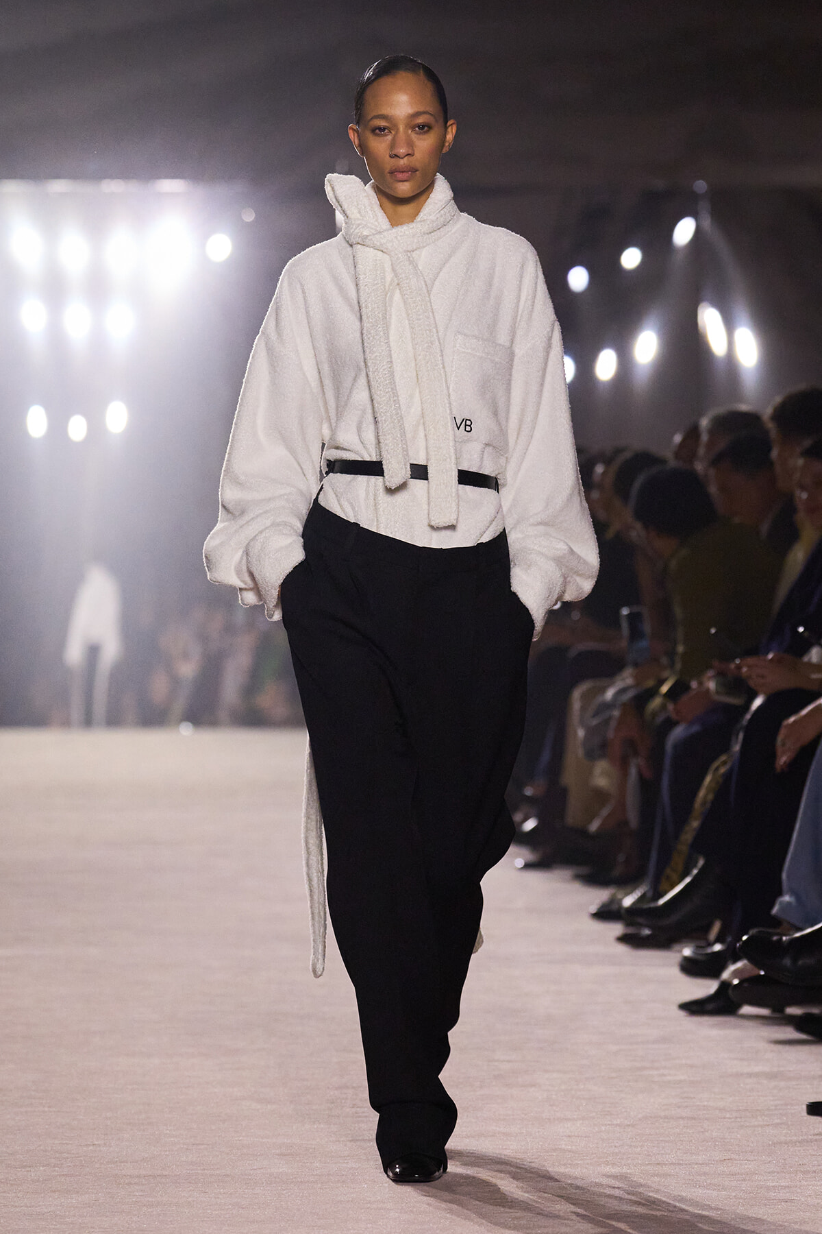 Runway model in an oversized white robe-like coat with a scarf tied at the neck, paired with black trousers; audience seated along the runway in the background.