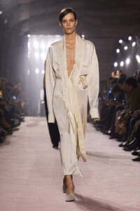Model walking a runway in a cream satin wrap robe with a belt and plunging neckline, wearing light gray heels.