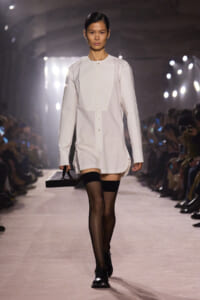 Model walking a runway in a white long-sleeve shirt dress with a concealed placket, carrying a black rectangular clutch, paired with sheer black tights and black oxford shoes.