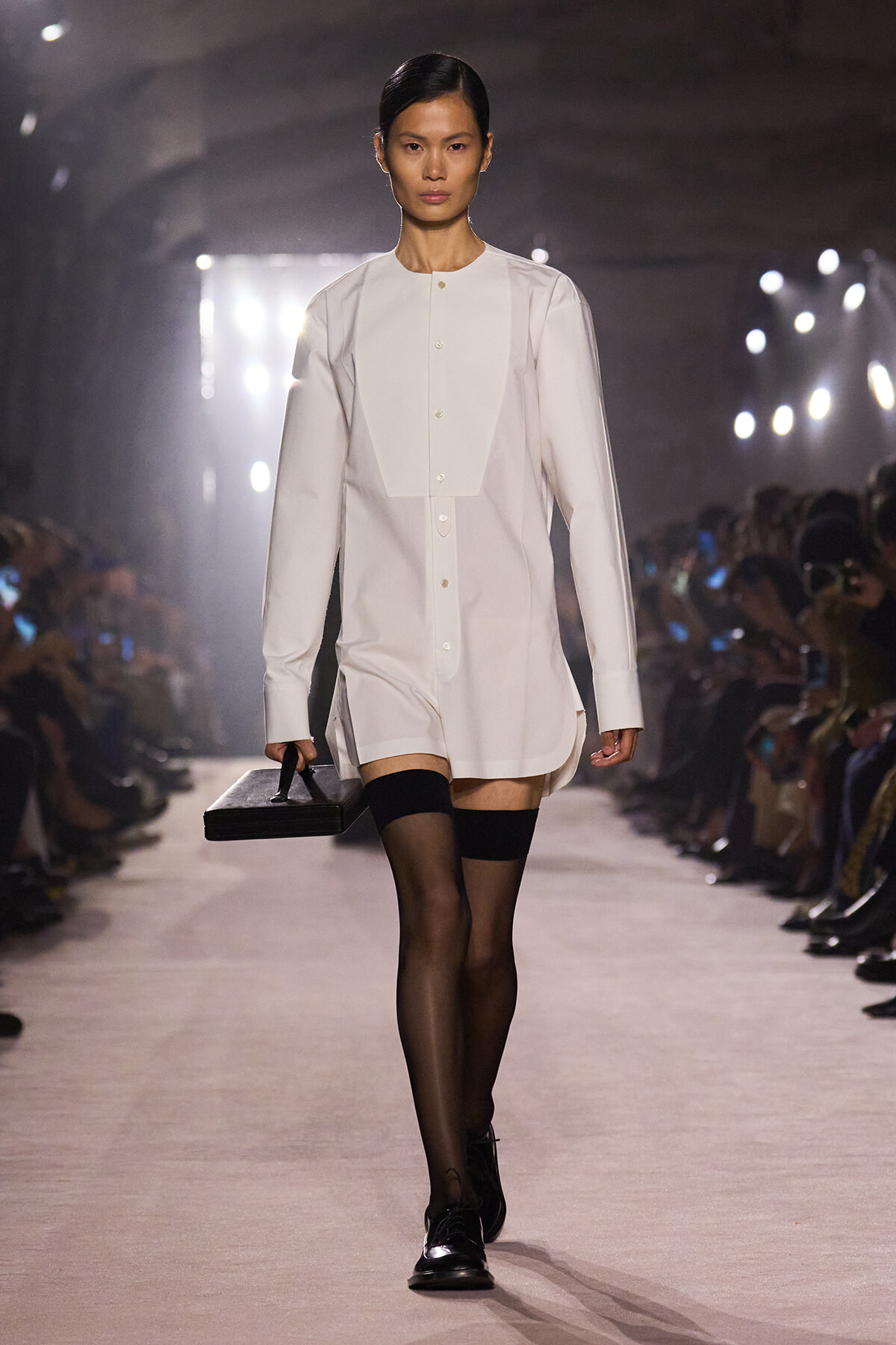 Model walking a runway in a white long-sleeve shirt dress with a concealed placket, carrying a black rectangular clutch, paired with sheer black tights and black oxford shoes.