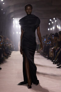 Model walks a dimly lit runway in a sleek, black satin gown with a high thigh-high slit and draped neckline.