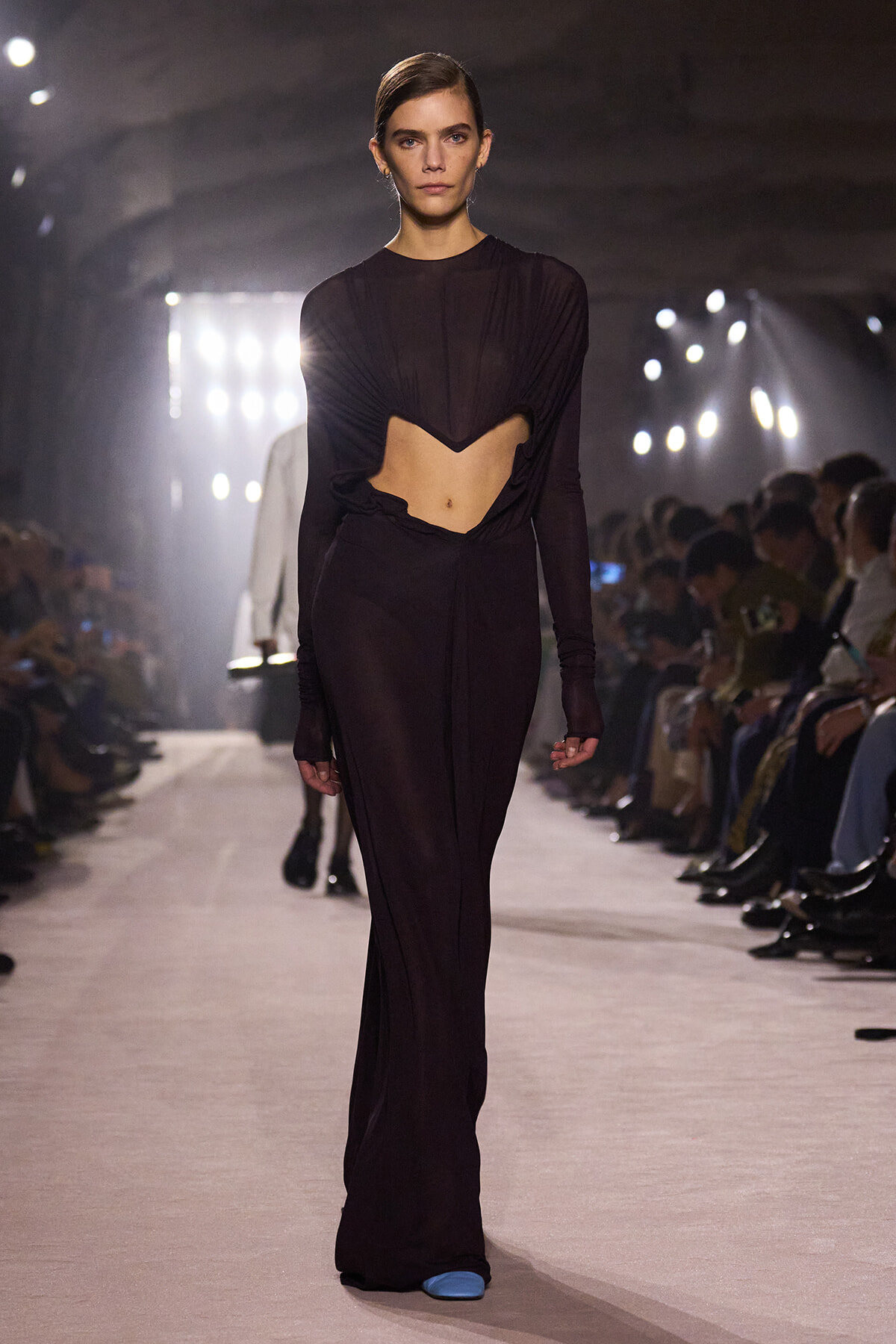 Model walks the runway in a burgundy long-sleeve crop top with a dramatic V-shaped midriff cutout and matching high-waisted pants; audience seated along the sides.