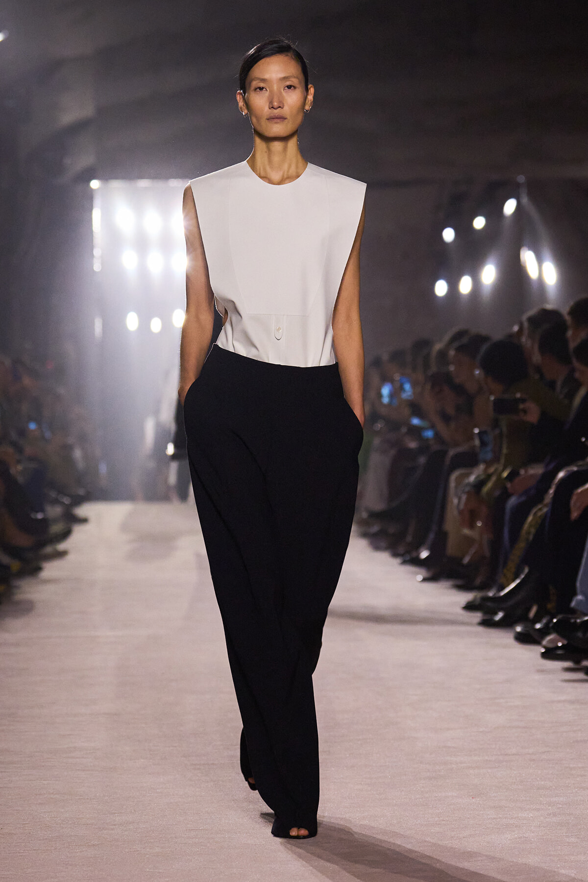 Model walking a runway in a white sleeveless top and black wide-leg pants; audience seated along the catwalk nearby.