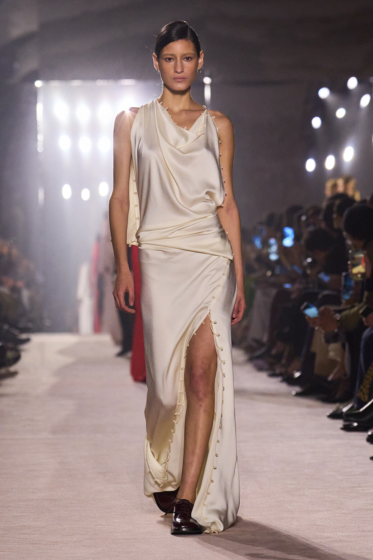 Model walking a fashion show wearing a cream draped satin gown with a high slit and lace-up detailing along the side, paired with dark loafers.