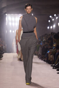 Model walks a runway in a sleeveless grey turtleneck knit top tucked into high-waisted grey trousers with a thin black belt; yellow shoes peek out.