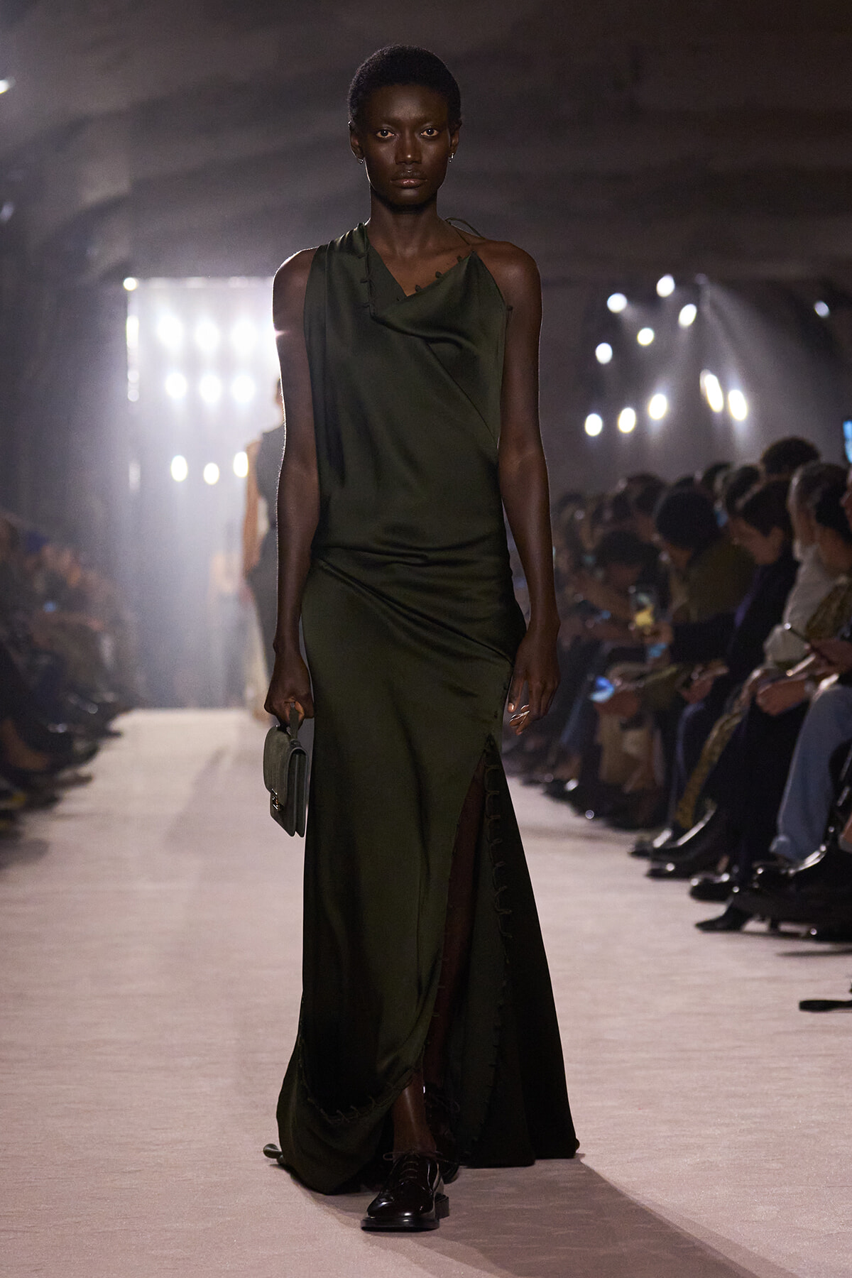 Model walks a runway in an olive-green draped one-shoulder gown with a high slit, holding a small clutch.