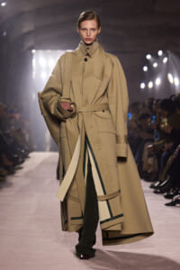 Model walks the runway in a tan oversized trench coat with a belted waist and wide cuffs, carrying a matching bag on her shoulder.