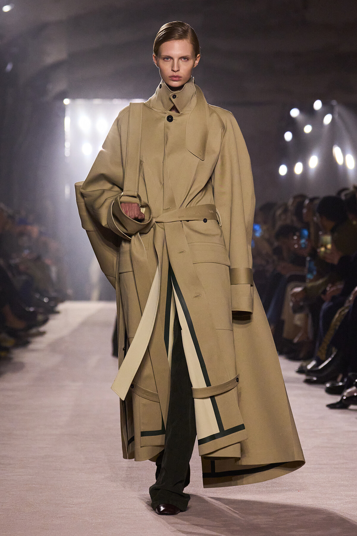 Model walks the runway in a tan oversized trench coat with a belted waist and wide cuffs, carrying a matching bag on her shoulder.