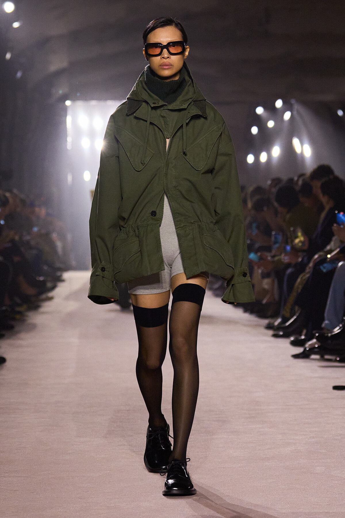 Model walks the runway in an oversized olive-green parka with large pockets, paired with gray shorts and black tights, wearing dark sunglasses and black shoes.