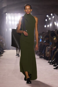 Model walking a runway in a dark olive green ribbed sleeveless dress with a high neck and asymmetrical scarf detail, carrying a large black tote bag over the shoulder and wearing black loafers.