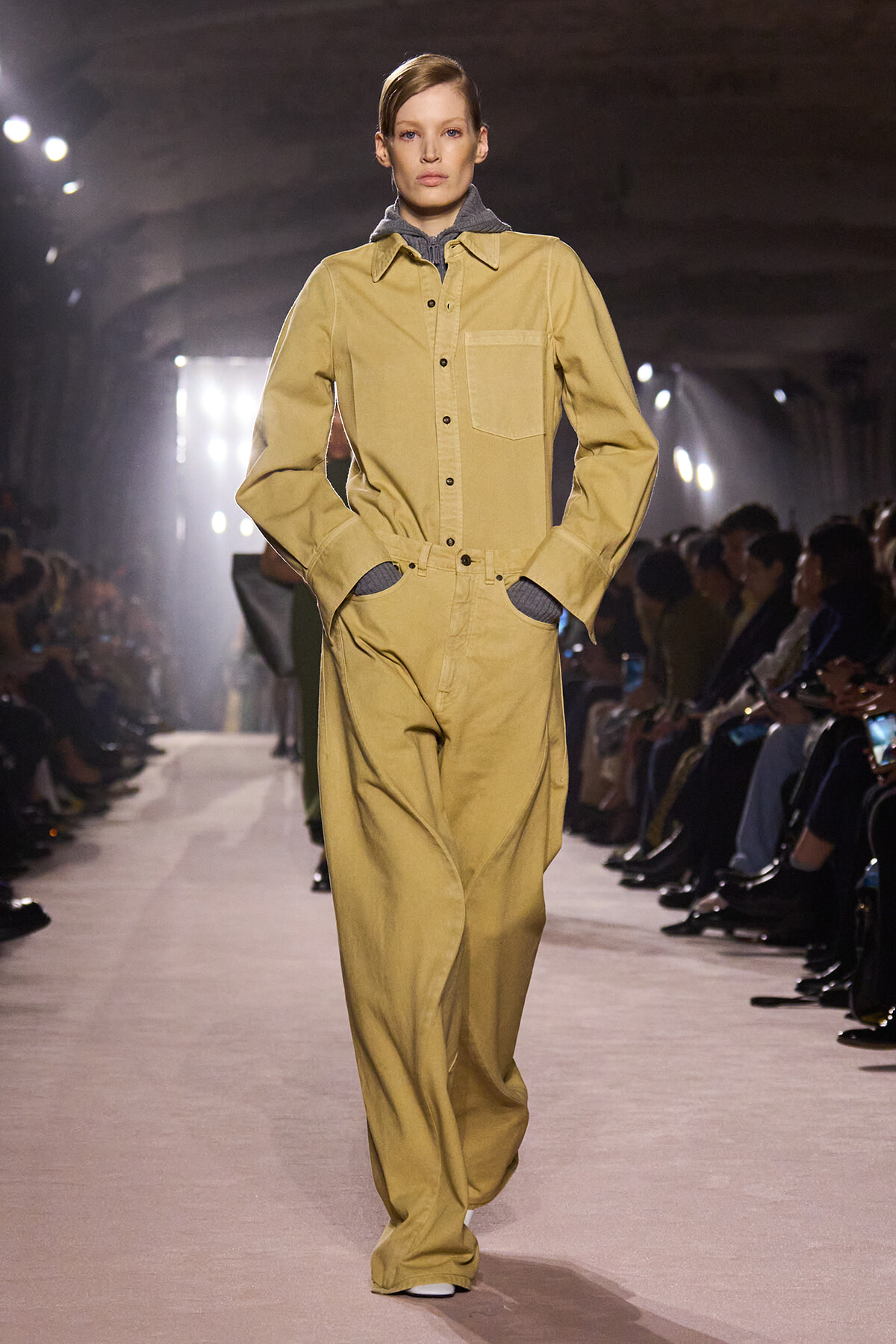 Model walking the runway in a tan utility jumpsuit with a chest pocket and wide legs, hands in pockets.
