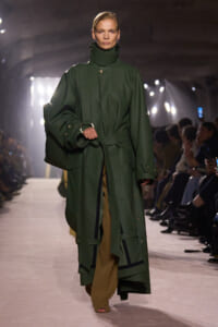 Model walking a runway in a long, dark green oversized trench coat with belt and high collar, tan pants peeking from underneath, audience along the sides.