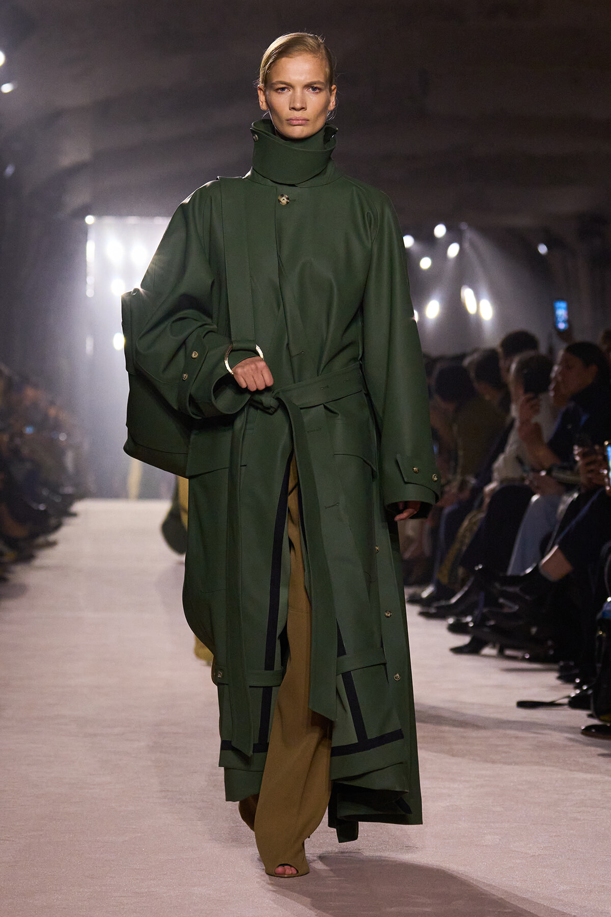 Model walking a runway in a long, dark green oversized trench coat with belt and high collar, tan pants peeking from underneath, audience along the sides.