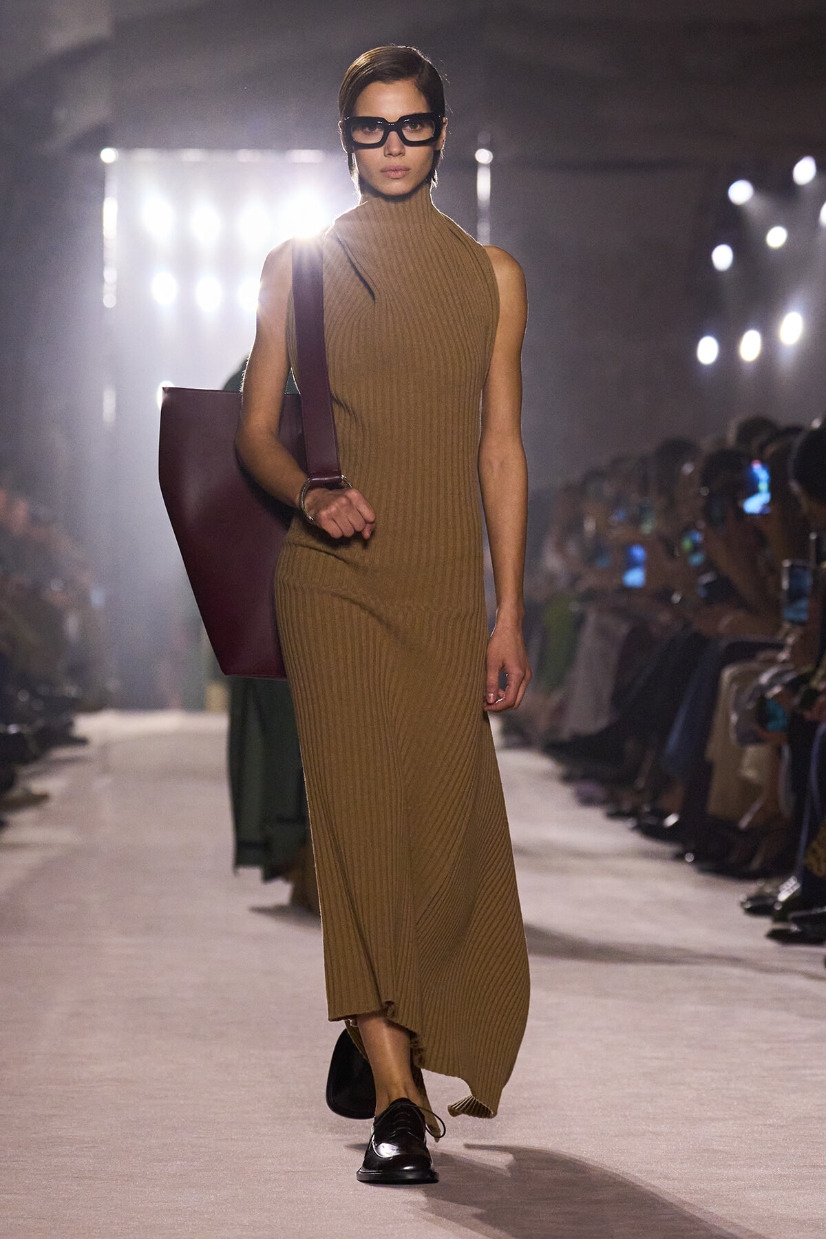 Model walking the runway in a camel ribbed sleeveless turtleneck maxi dress, black sunglasses, and a large burgundy tote bag.