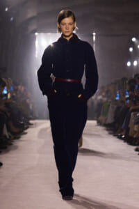 Model walks a fashion runway in a navy jumpsuit with a brown belt, hands in pockets, audience visible on both sides.