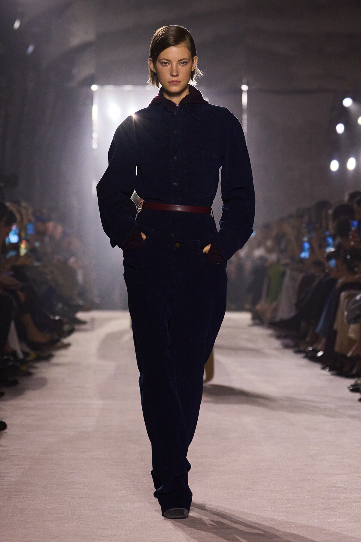 Model walks a fashion runway in a navy jumpsuit with a brown belt, hands in pockets, audience visible on both sides.