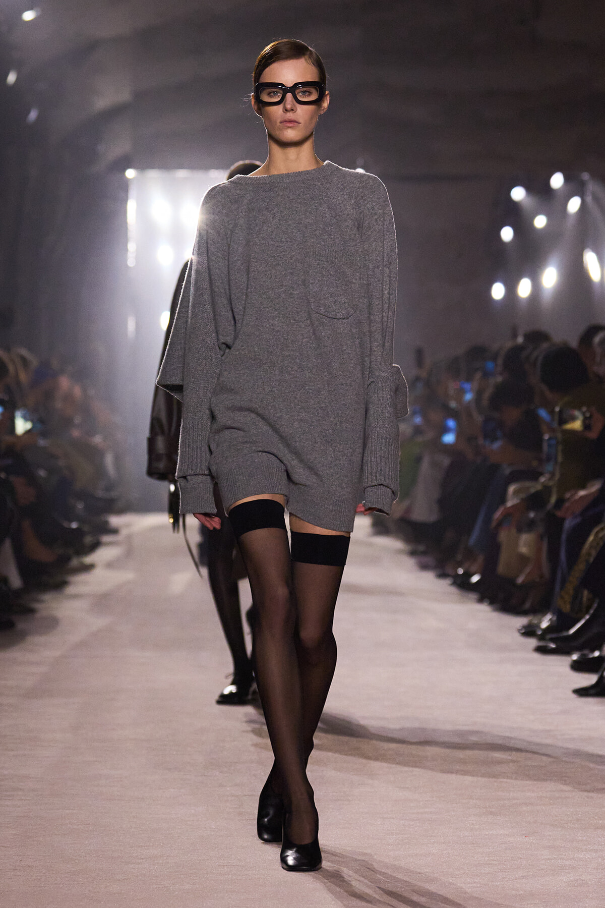 Model walking a fashion runway in an oversized gray sweater-dress, black tights, and black heels, wearing thick black glasses.