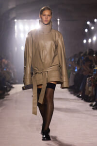 Model on a fashion runway wearing a beige oversized leather coat with a wide belt, black stockings, and black shoes.
